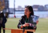 On East Boston’s waterfront, Mayor Wu releases 2030 Climate Action Plan Asian woman speaking at a wooden podium outdoors with a microphone, holding a pamphlet titled '2030 Climate Action Plan' as city skyline and water appear in the background