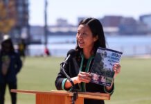 On East Boston’s waterfront, Mayor Wu releases 2030 Climate Action Plan Asian woman speaking at a wooden podium outdoors with a microphone, holding a pamphlet titled '2030 Climate Action Plan' as city skyline and water appear in the background