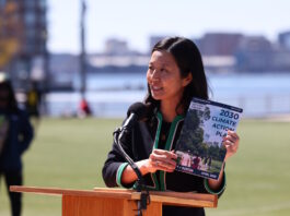 On East Boston’s waterfront, Mayor Wu releases 2030 Climate Action Plan Asian woman speaking at a wooden podium outdoors with a microphone, holding a pamphlet titled '2030 Climate Action Plan' as city skyline and water appear in the background