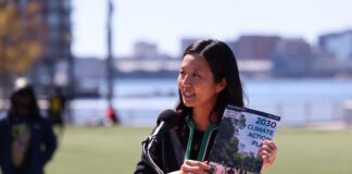 On East Boston’s waterfront, Mayor Wu releases 2030 Climate Action Plan Asian woman speaking at a wooden podium outdoors with a microphone, holding a pamphlet titled '2030 Climate Action Plan' as city skyline and water appear in the background