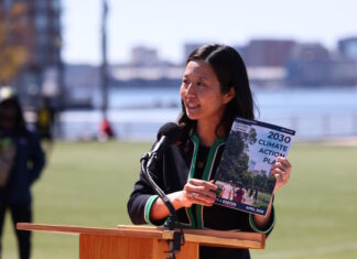 On East Boston’s waterfront, Mayor Wu releases 2030 Climate Action Plan Asian woman speaking at a wooden podium outdoors with a microphone, holding a pamphlet titled '2030 Climate Action Plan' as city skyline and water appear in the background