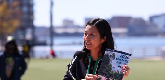 On East Boston’s waterfront, Mayor Wu releases 2030 Climate Action Plan Asian woman speaking at a wooden podium outdoors with a microphone, holding a pamphlet titled '2030 Climate Action Plan' as city skyline and water appear in the background