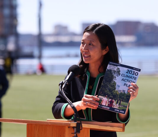 On East Boston’s waterfront, Mayor Wu releases 2030 Climate Action Plan Asian woman speaking at a wooden podium outdoors with a microphone, holding a pamphlet titled '2030 Climate Action Plan' as city skyline and water appear in the background