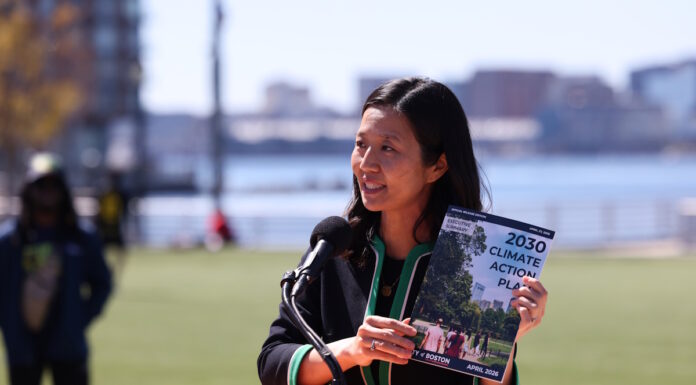 On East Boston’s waterfront, Mayor Wu releases 2030 Climate Action Plan Asian woman speaking at a wooden podium outdoors with a microphone, holding a pamphlet titled '2030 Climate Action Plan' as city skyline and water appear in the background