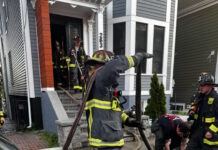 Social Centers establishes fund for victims of Princeton Street fire Firefighters in turnout gear handling large hoses on a residential street, preparing entry into a house with crews in the doorway.