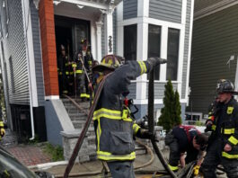 Social Centers establishes fund for victims of Princeton Street fire Firefighters in turnout gear handling large hoses on a residential street, preparing entry into a house with crews in the doorway.