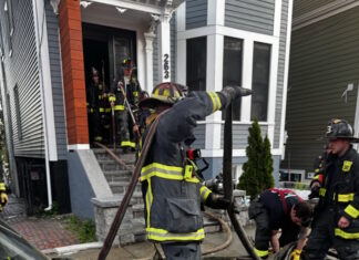 Social Centers establishes fund for victims of Princeton Street fire Firefighters in turnout gear handling large hoses on a residential street, preparing entry into a house with crews in the doorway.