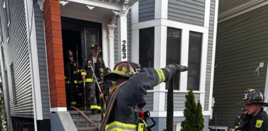Social Centers establishes fund for victims of Princeton Street fire Firefighters in turnout gear handling large hoses on a residential street, preparing entry into a house with crews in the doorway.