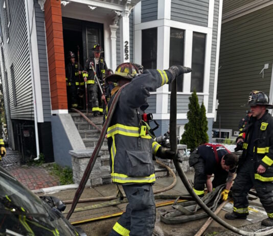 Social Centers establishes fund for victims of Princeton Street fire Firefighters in turnout gear handling large hoses on a residential street, preparing entry into a house with crews in the doorway.