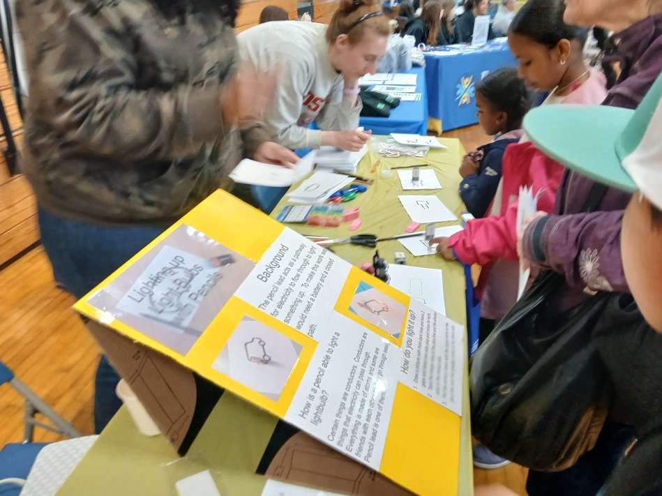 People gathered around a table at a community fair, working on a yellow project board with diagrams and text.","Volunteer helps children with crafts as they read from a large yellow informational board.","Children and adults fill out papers and cut shapes at a booth featuring a bright yellow display panel.","Hands-on activity at a booth; the yellow project board displays images and written info.