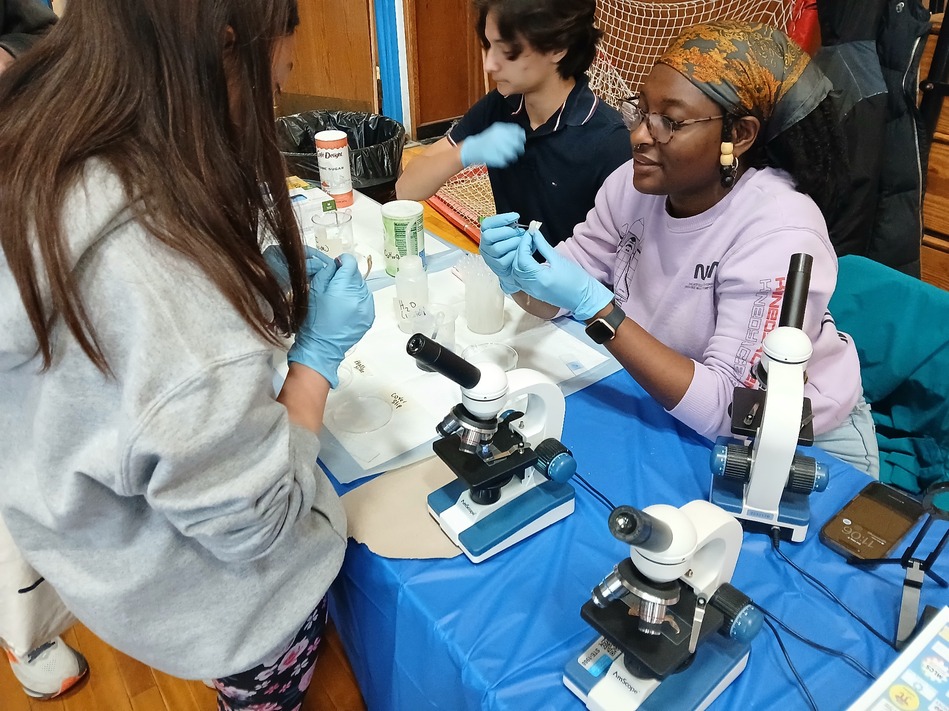 Group of people in gloves conducting a hands-on science experiment at a table with microscopes and lab gear.
