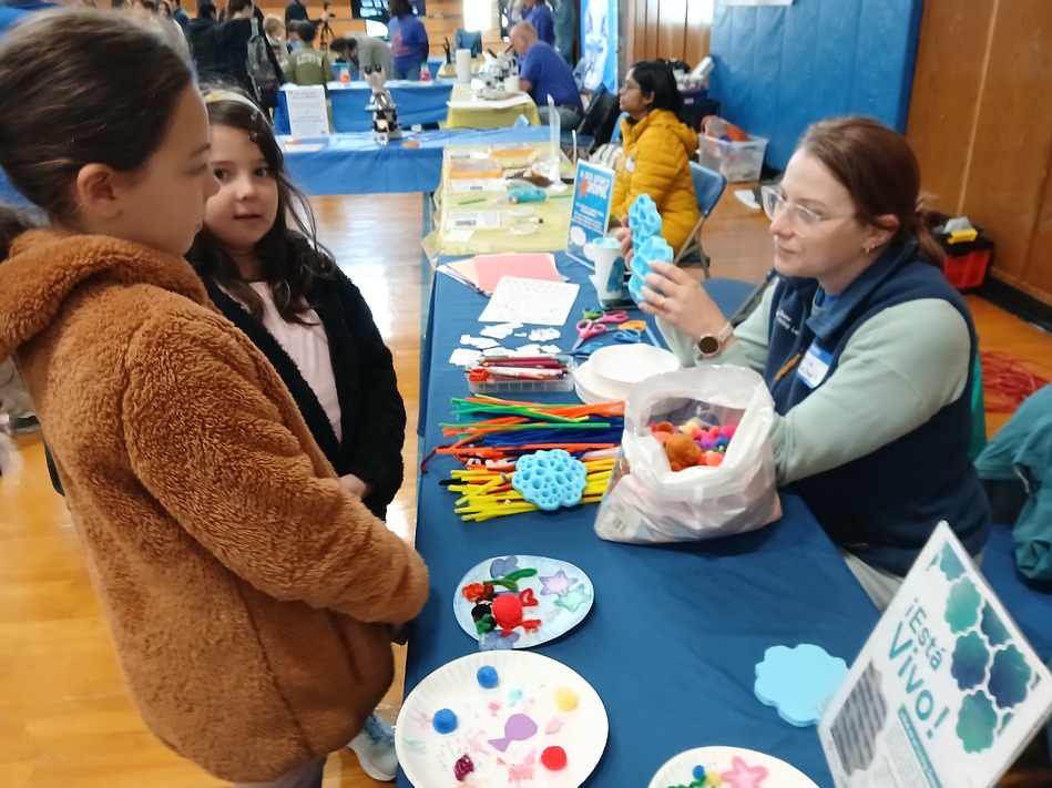Two young girls watch a staff member at a craft table during a community event, with colorful craft supplies like foam shapes and pipe cleaners on the table.