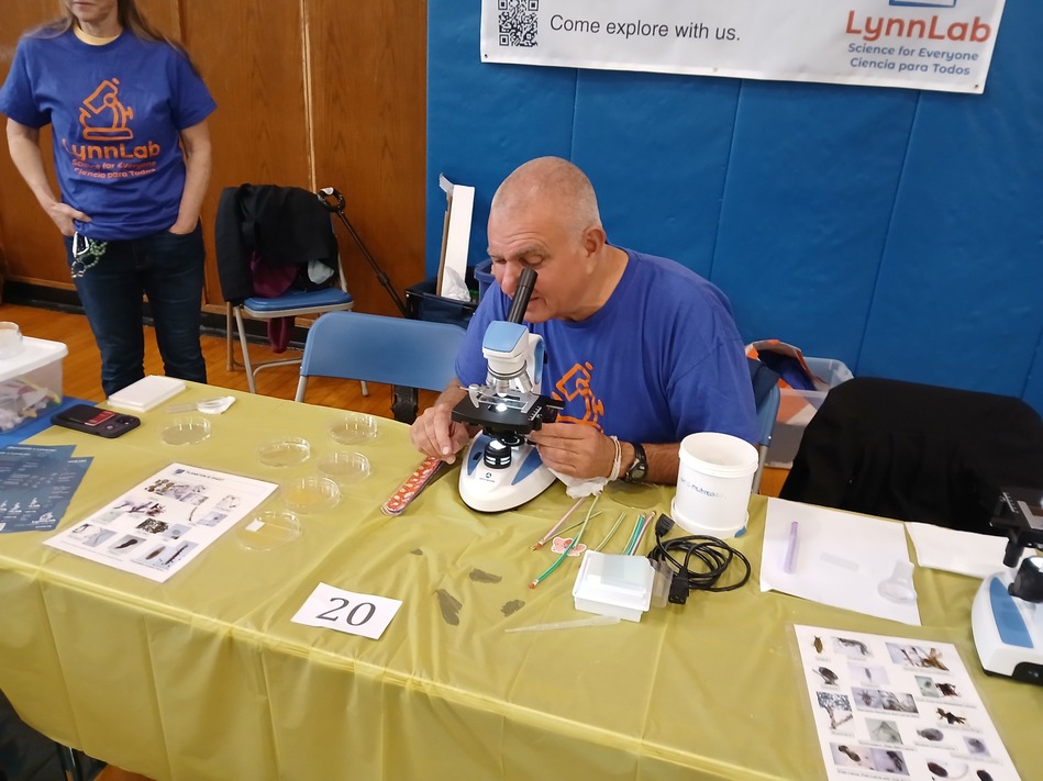 Man in a blue LynnLab shirt using a microscope at a science booth, with a woman standing nearby and lab materials on the table behind a yellow cloth.