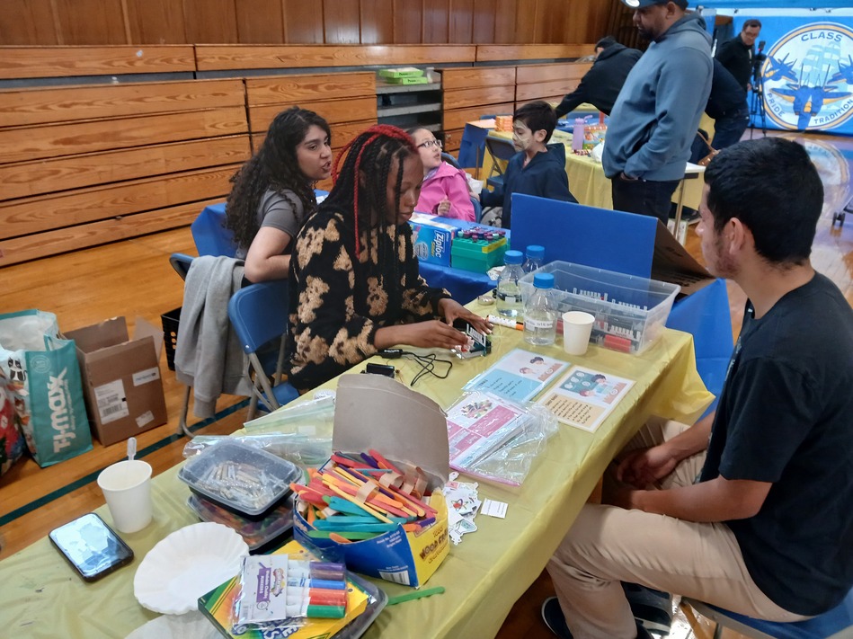 Group of diverse teens and young adults seated around a table in a gym, doing crafts with markers and supplies; volunteers help at a community event.