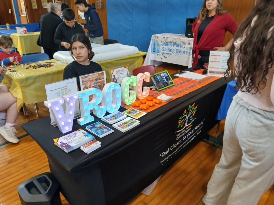 Community event booth with illuminated letters spelling VROGCE on a table, brochures and a tablet on display