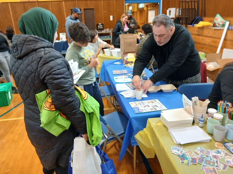 Volunteer helps children with a craft activity at a community fair table in a gymnasium.