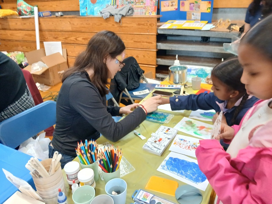 Instructor helps a child with a craft at a colorful art table, surrounded by paints, brushes, and drawings in a classroom setting.