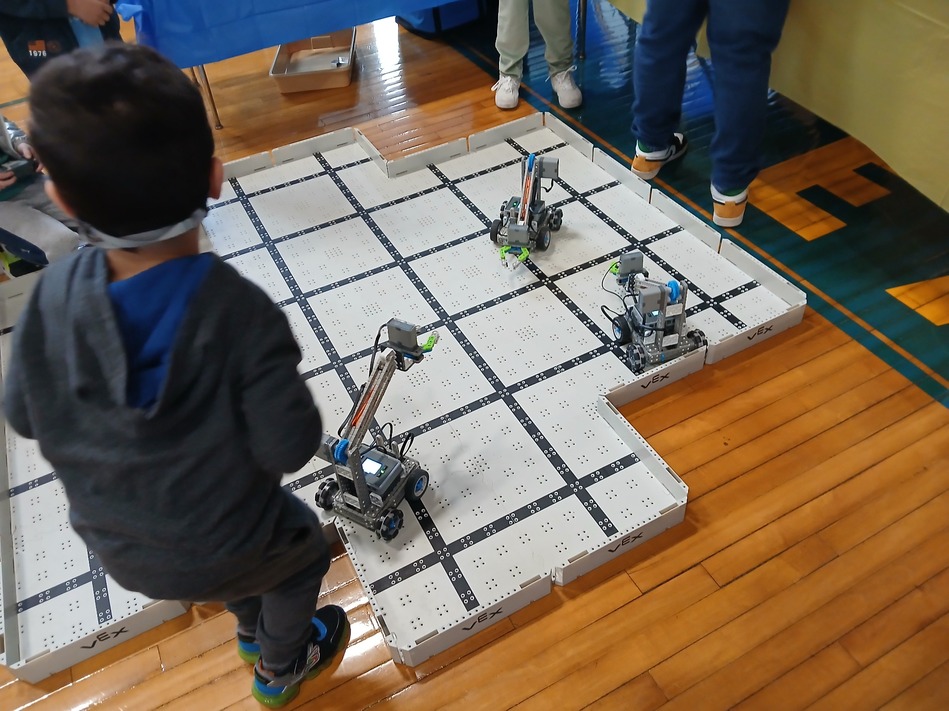 Children watching a white grid robotics arena in a gym as small VEX robots navigate the squares.