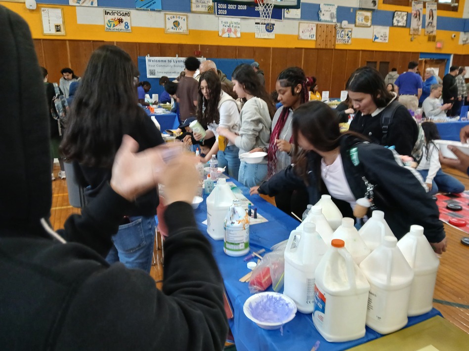 Group of students around a long table with gallon jugs and bowls in a gym during a community science fair or expo.