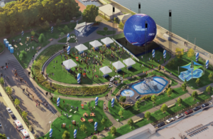 Aerial view of a waterfront festival with white tents, a circular lawn, a large blue inflatable stage, seating areas, and a water feature.