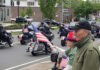 Set the date: 5/17 — 15th Annual Wounded Veterans Run Motorcycle parade on a city street; riders with American flags pass by while spectators on the curb wave flags.