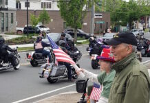 Set the date: 5/17 — 15th Annual Wounded Veterans Run Motorcycle parade on a city street; riders with American flags pass by while spectators on the curb wave flags.