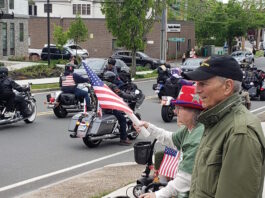 Set the date: 5/17 — 15th Annual Wounded Veterans Run Motorcycle parade on a city street; riders with American flags pass by while spectators on the curb wave flags.