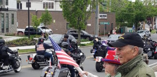 Set the date: 5/17 — 15th Annual Wounded Veterans Run Motorcycle parade on a city street; riders with American flags pass by while spectators on the curb wave flags.