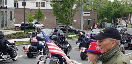 Set the date: 5/17 — 15th Annual Wounded Veterans Run Motorcycle parade on a city street; riders with American flags pass by while spectators on the curb wave flags.