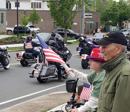 Set the date: 5/17 — 15th Annual Wounded Veterans Run Motorcycle parade on a city street; riders with American flags pass by while spectators on the curb wave flags.