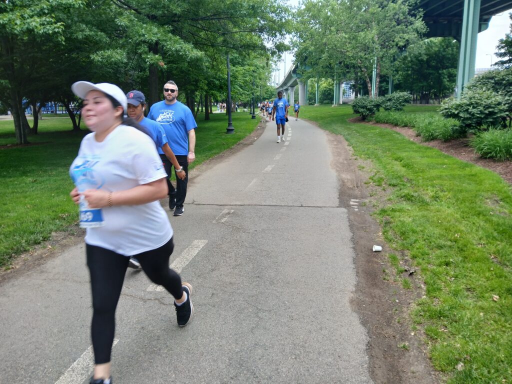 Foreground runner in a white cap leads a group of joggers in blue shirts along a tree-lined park path.