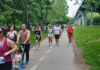 NeigborHealth’s Get Movin’ run a success as hundreds of runners and walkers promote health and wellness People walking/running on a park trail with bib numbers; a man pushes a blue stroller with a child in front, trees and a bridge in the background.