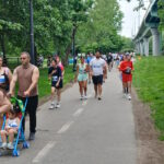 NeigborHealth’s Get Movin’ run a success as hundreds of runners and walkers promote health and wellness People walking/running on a park trail with bib numbers; a man pushes a blue stroller with a child in front, trees and a bridge in the background.