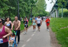 NeigborHealth’s Get Movin’ run a success as hundreds of runners and walkers promote health and wellness People walking/running on a park trail with bib numbers; a man pushes a blue stroller with a child in front, trees and a bridge in the background.