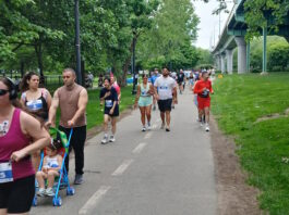 NeigborHealth’s Get Movin’ run a success as hundreds of runners and walkers promote health and wellness People walking/running on a park trail with bib numbers; a man pushes a blue stroller with a child in front, trees and a bridge in the background.