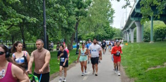 NeigborHealth’s Get Movin’ run a success as hundreds of runners and walkers promote health and wellness People walking/running on a park trail with bib numbers; a man pushes a blue stroller with a child in front, trees and a bridge in the background.