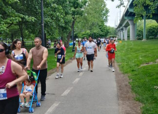 NeigborHealth’s Get Movin’ run a success as hundreds of runners and walkers promote health and wellness People walking/running on a park trail with bib numbers; a man pushes a blue stroller with a child in front, trees and a bridge in the background.