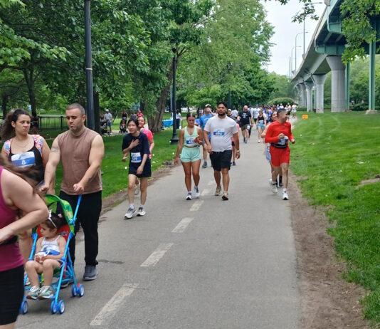 NeigborHealth’s Get Movin’ run a success as hundreds of runners and walkers promote health and wellness People walking/running on a park trail with bib numbers; a man pushes a blue stroller with a child in front, trees and a bridge in the background.