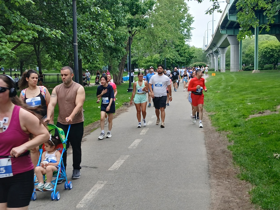 People walking/running on a park trail with bib numbers; a man pushes a blue stroller with a child in front, trees and a bridge in the background.