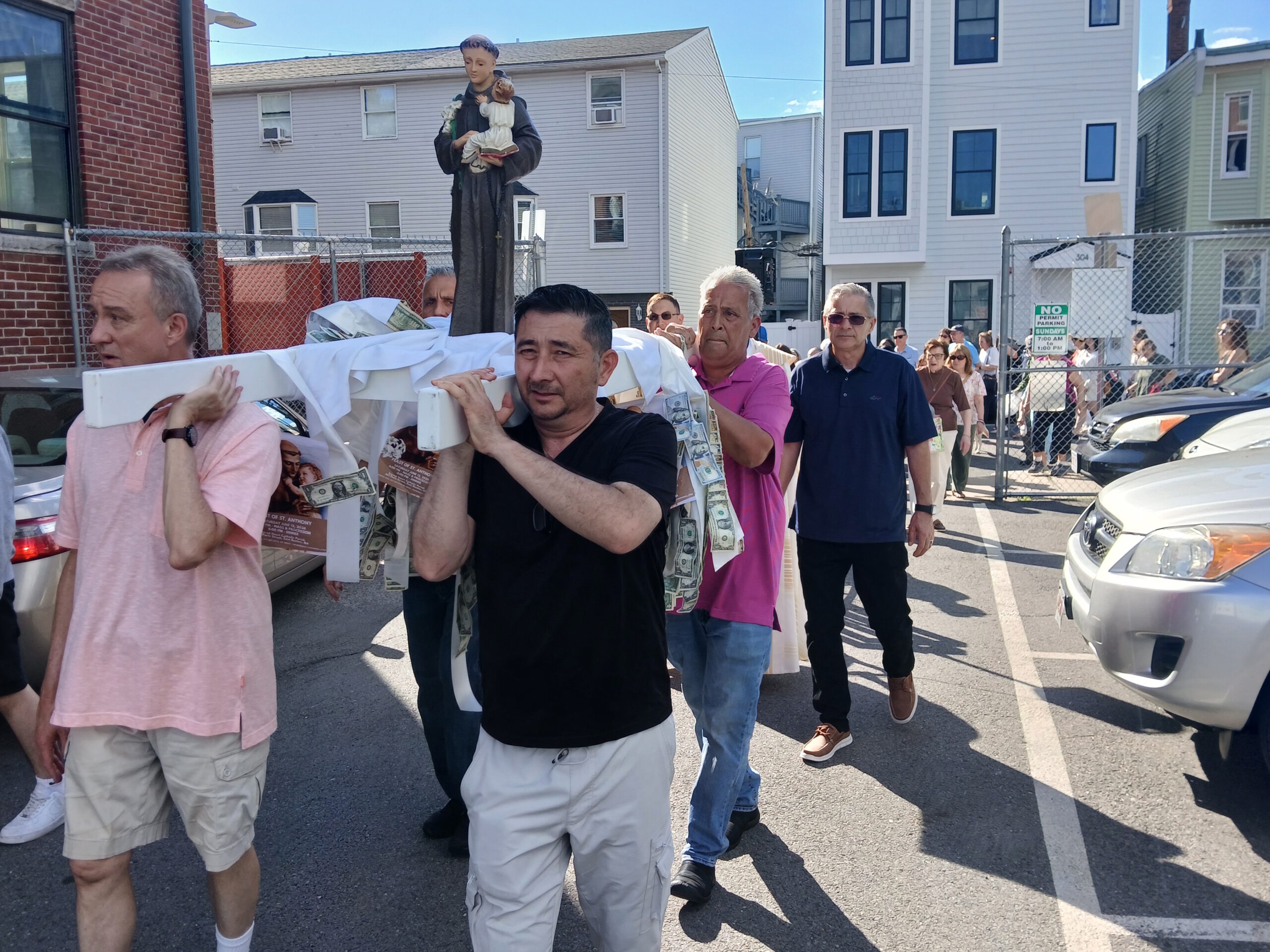 A group of men carrying a religious float with a statue of a saint holding a child, outdoors on a sunny street, with dollar bills taped to the float and onlookers in the background.