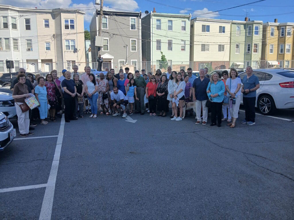 Large diverse group poses for a group photo in a parking lot, with colorful row houses in the background.