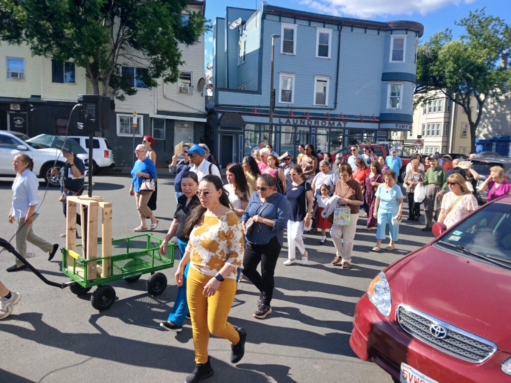 Crowd of people walking in a street parade past a blue laundromat building, with a green wagon in the foreground and a red car on the right side.
