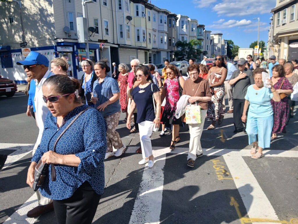 Crowd of mostly older adults walking together on a city street during a parade, with row of houses and storefronts in the background.