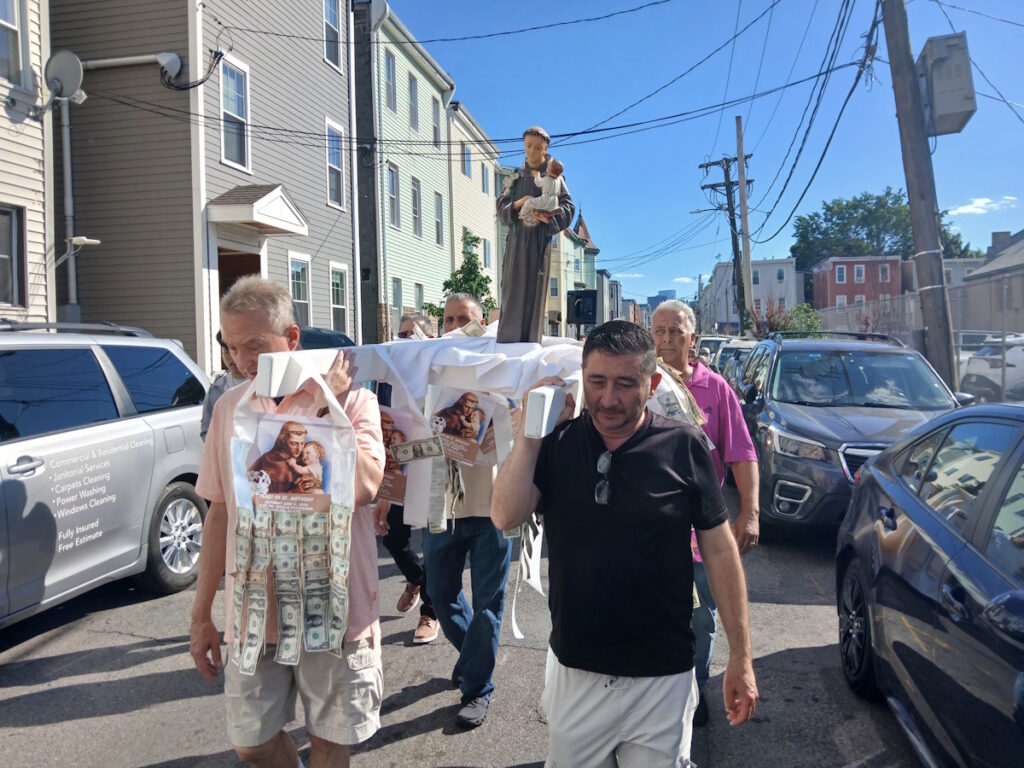 Religious street procession with a saint statue carried on a white-draped platform along a neighborhood street.