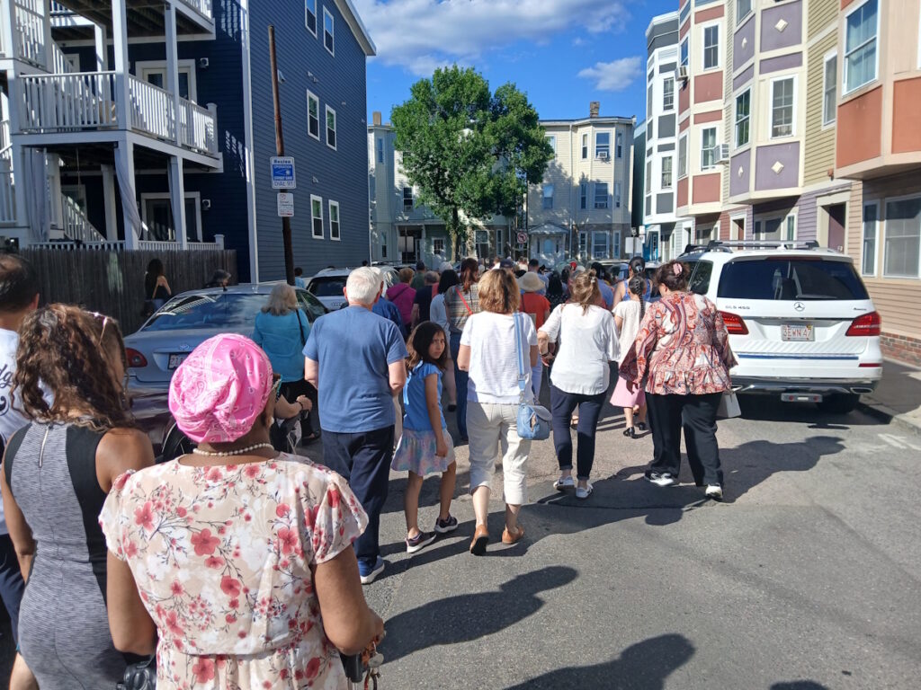 Crowd of people walking down a sunlit residential street between colorful apartment buildings; parked cars along the curb and a clear blue sky.