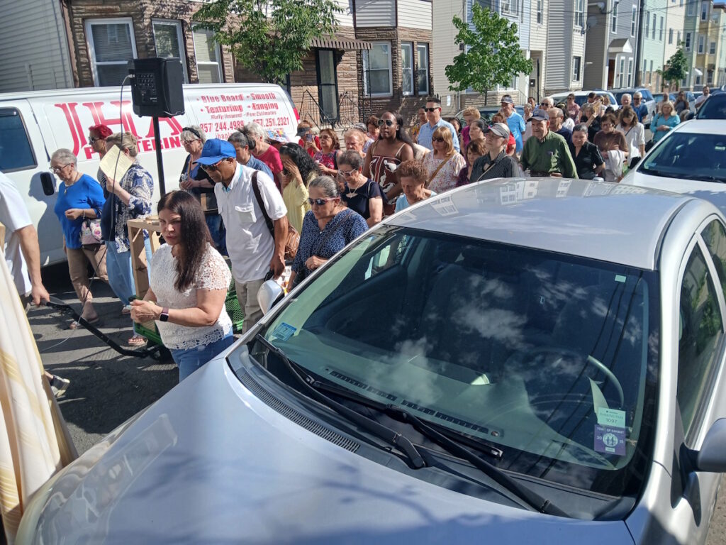 A crowd of people walking down a residential street in daytime next to a white van with red lettering, with a speaker on a stand nearby and cars parked along the curb, suggesting a street procession or event.