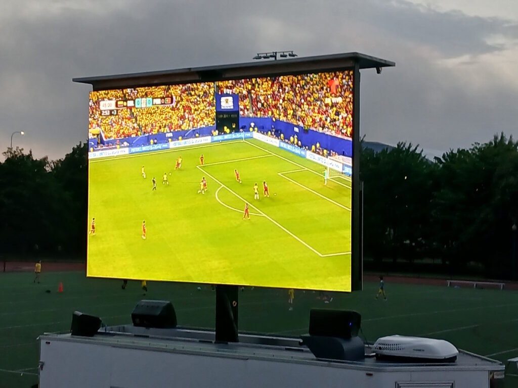 Large outdoor stadium screen showing a live soccer match with players on a green pitch and a cheering crowd in the stands behind the goal.