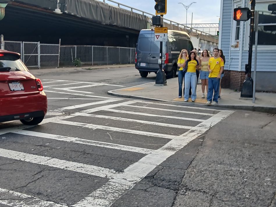Group of five people in yellow shirts waiting to cross a city street at a zebra crosswalk.