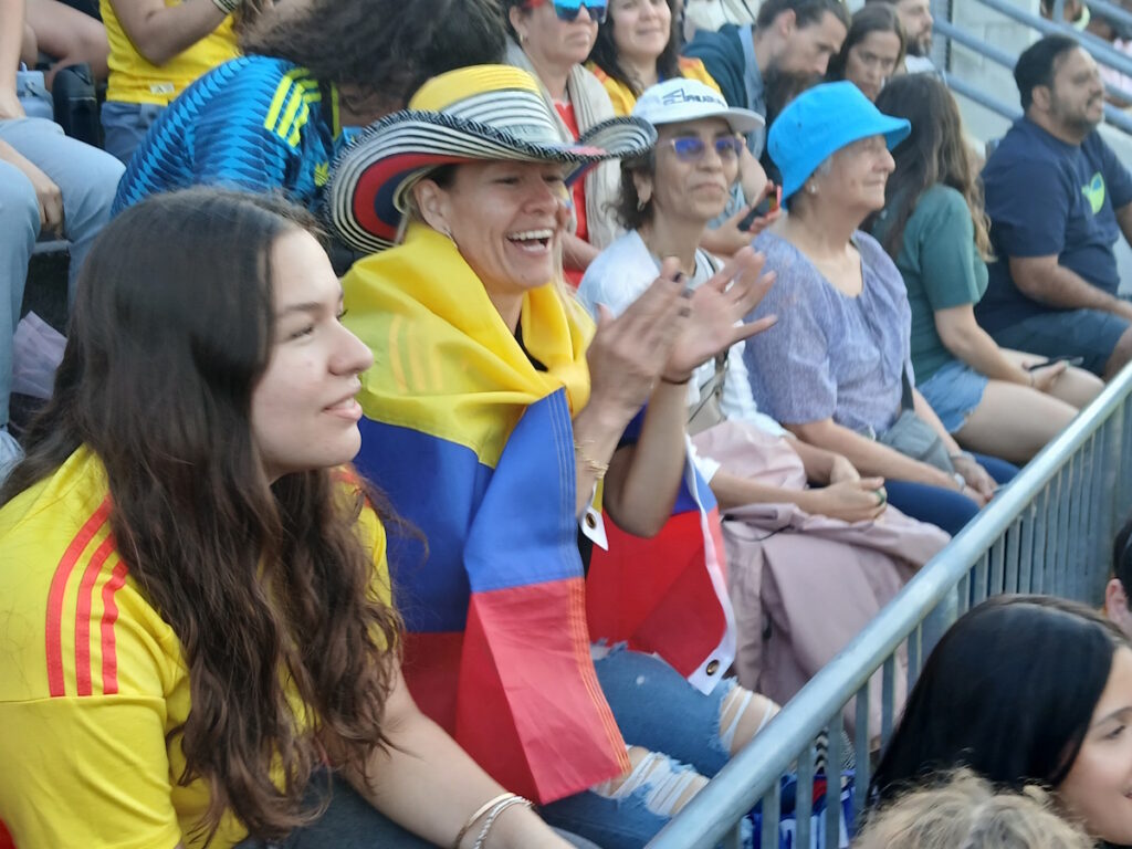 Spectators at a stadium, a woman with a striped hat and a Colombian flag draped over her shoulders, clapping.