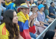 Scenes from the Columbia v. Portugal watch party at East Boston Stadium Spectators at a stadium, a woman with a striped hat and a Colombian flag draped over her shoulders, clapping.